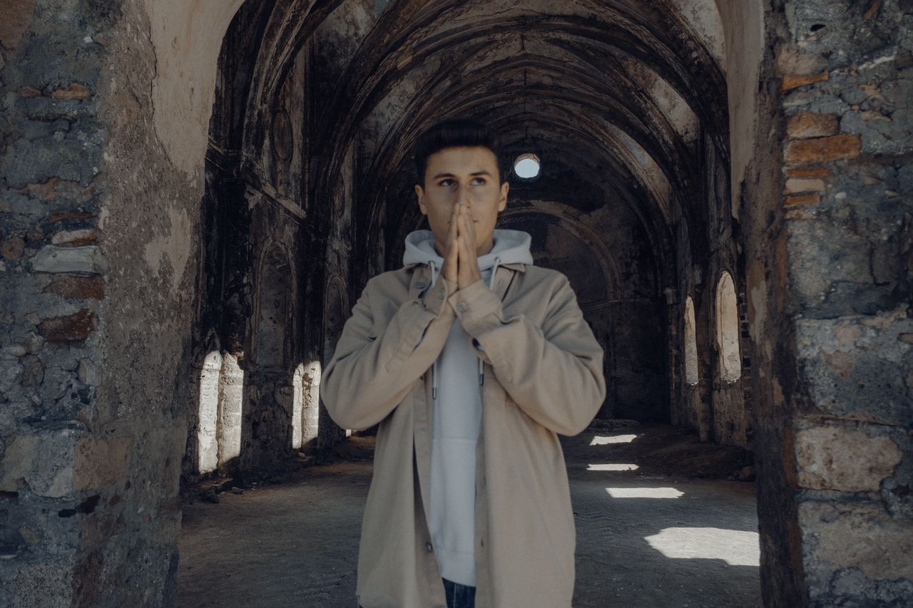 a young man in a hoodie poses thoughtfully in a historic stone arcade