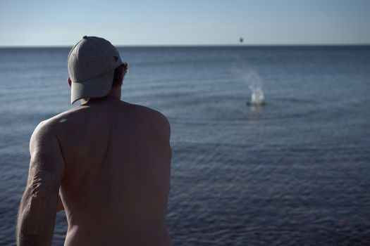A shirtless man in a cap throws stones into the sea on a sunny summer day.