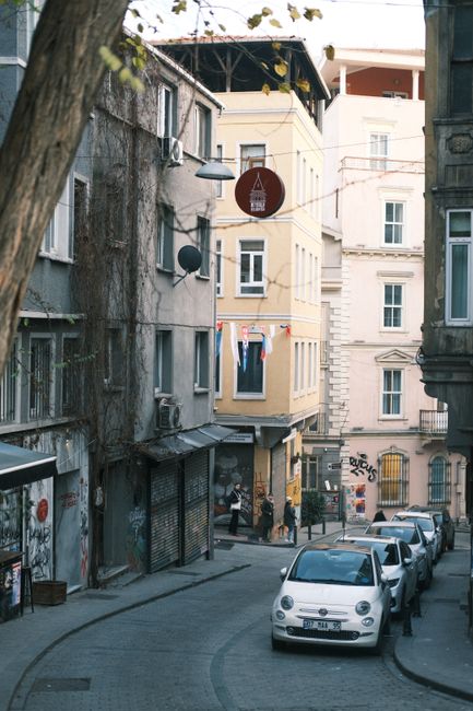 Narrow city street lined with historic buildings, showcasing urban life with parked cars and pedestrians.