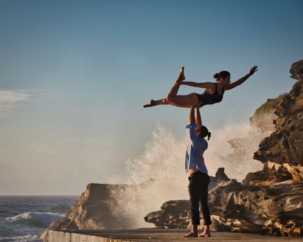 A man and woman perform an acro yoga pose on a rocky shore with waves crashing at Tamarama Beach, Australia.