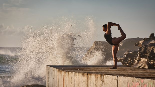 Woman practicing yoga on concrete pier with dramatic ocean waves at Tamarama Beach, Australia.