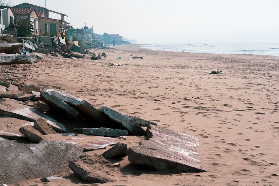 Photo by Theologos Christodoulou A serene coastal scene with broken concrete along a sandy shoreline and homes in the background.