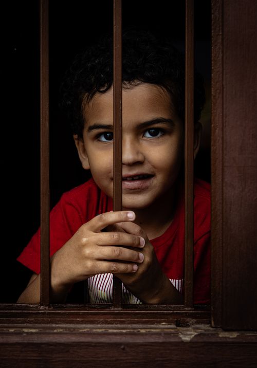 Free Portrait of a cheerful child smiling while peeking through a barred window indoors. Stock Photo