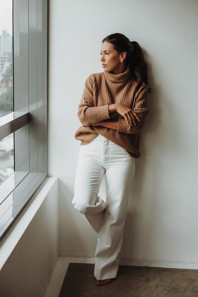 Woman In White Pants Leaning Against A Wall And Looking Out The Window