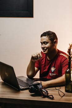 A smiling man in a casual red shirt works on a laptop at a dimly lit desk.