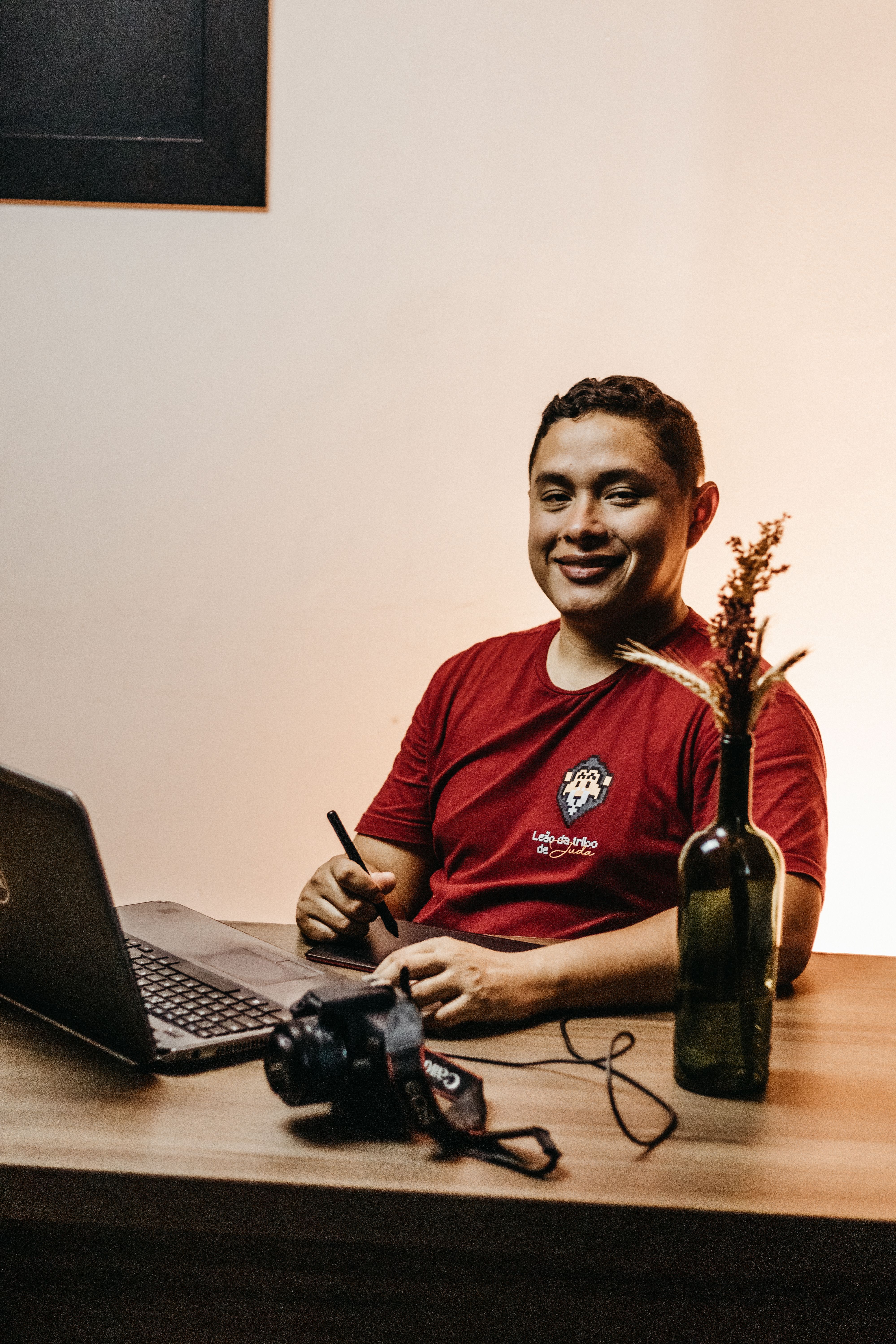 A smiling man sitting at a desk working on a laptop with a camera and dried flowers in a vase.