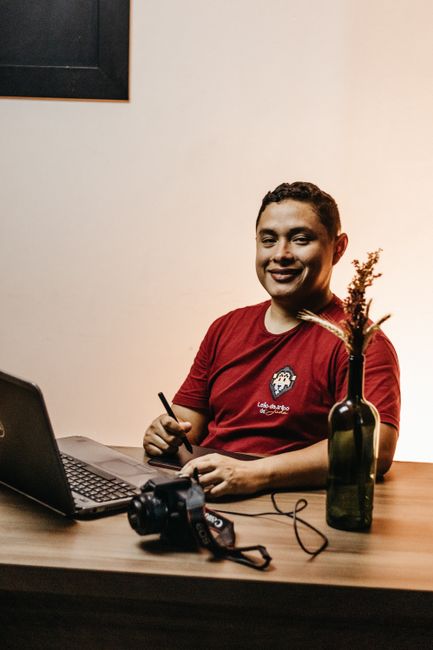 A smiling man sitting at a desk working on a laptop with a camera and dried flowers in a vase. A smiling man sitting at a desk working on a laptop with a camera and dried flowers in a vase.
