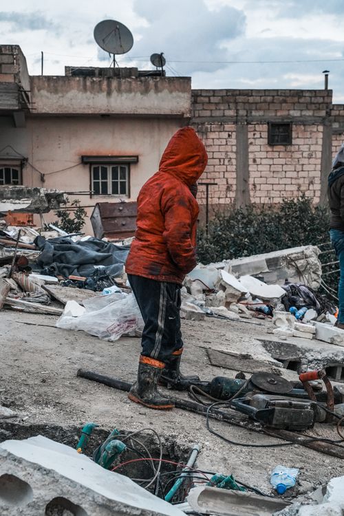 Free A child in a red jacket stands among debris in Jindires, Syria, reflecting urban devastation. Stock Photo