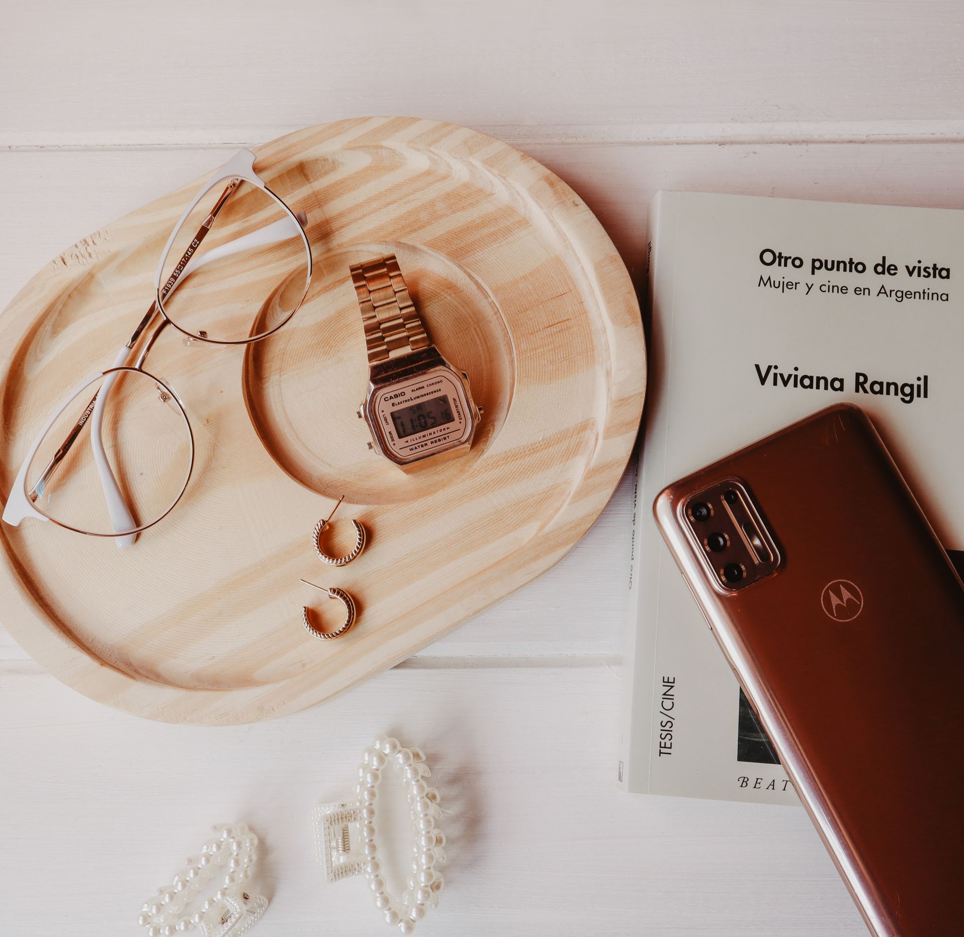 Elegant flat lay of accessories including a gold watch, glasses, earrings, and a smartphone on a wooden tray with a book.