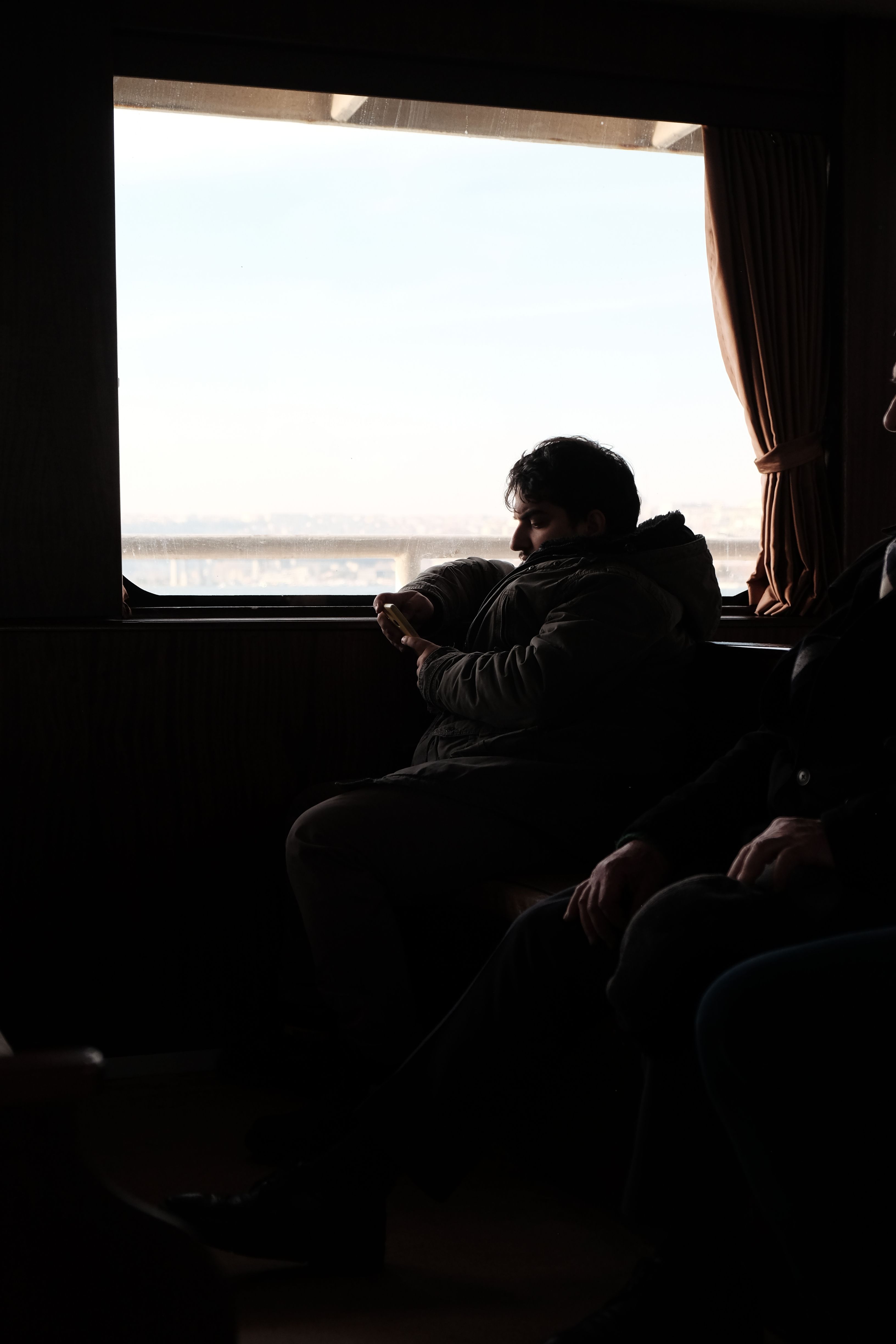 A man in a jacket sits by a ferry window, lost in thought, with daylight streaming in.