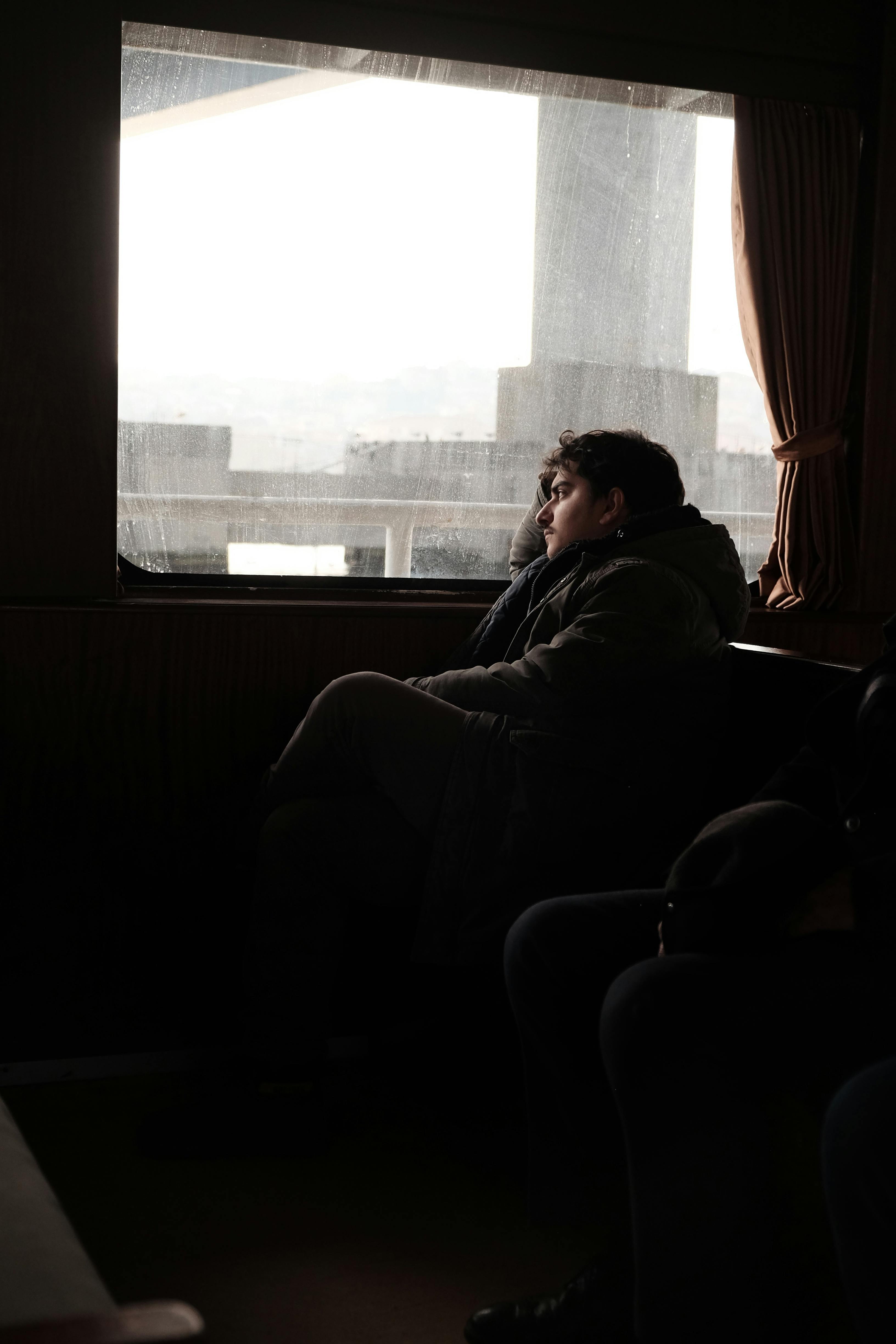 A man in a jacket sits on a ferry, looking thoughtfully out the window as the scenery passes by.
