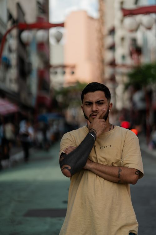 Free Contemplative man with tattoos stands on a bustling city street, embodying modern urban lifestyle. Stock Photo