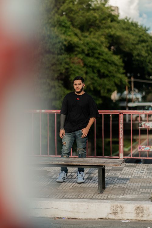 Free A man in casual attire standing against an urban bridge railing with trees in the background. Stock Photo