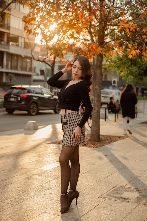 Young Fashionable Woman in a Skirt and Heels Standing on a Sidewalk on the Background of Autumnal Trees