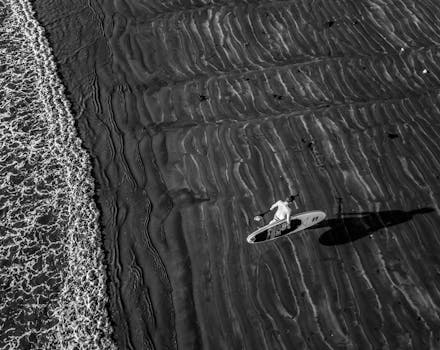 Black and white aerial shot of a surfer carrying a board on a textured sandy beach.