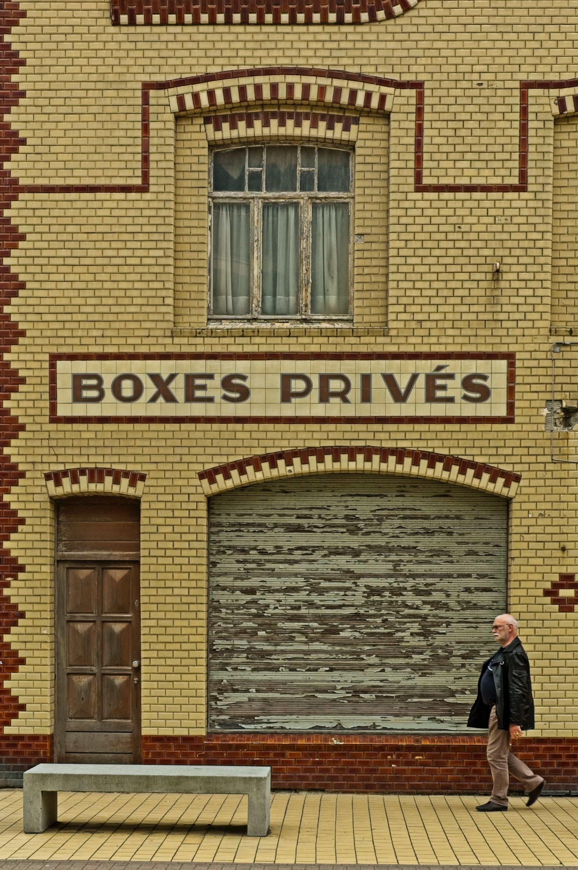 Free Yellow brick facade with 'BOXES PRIVÉS' sign and a man walking by in Coxyde, Belgium. Stock Photo