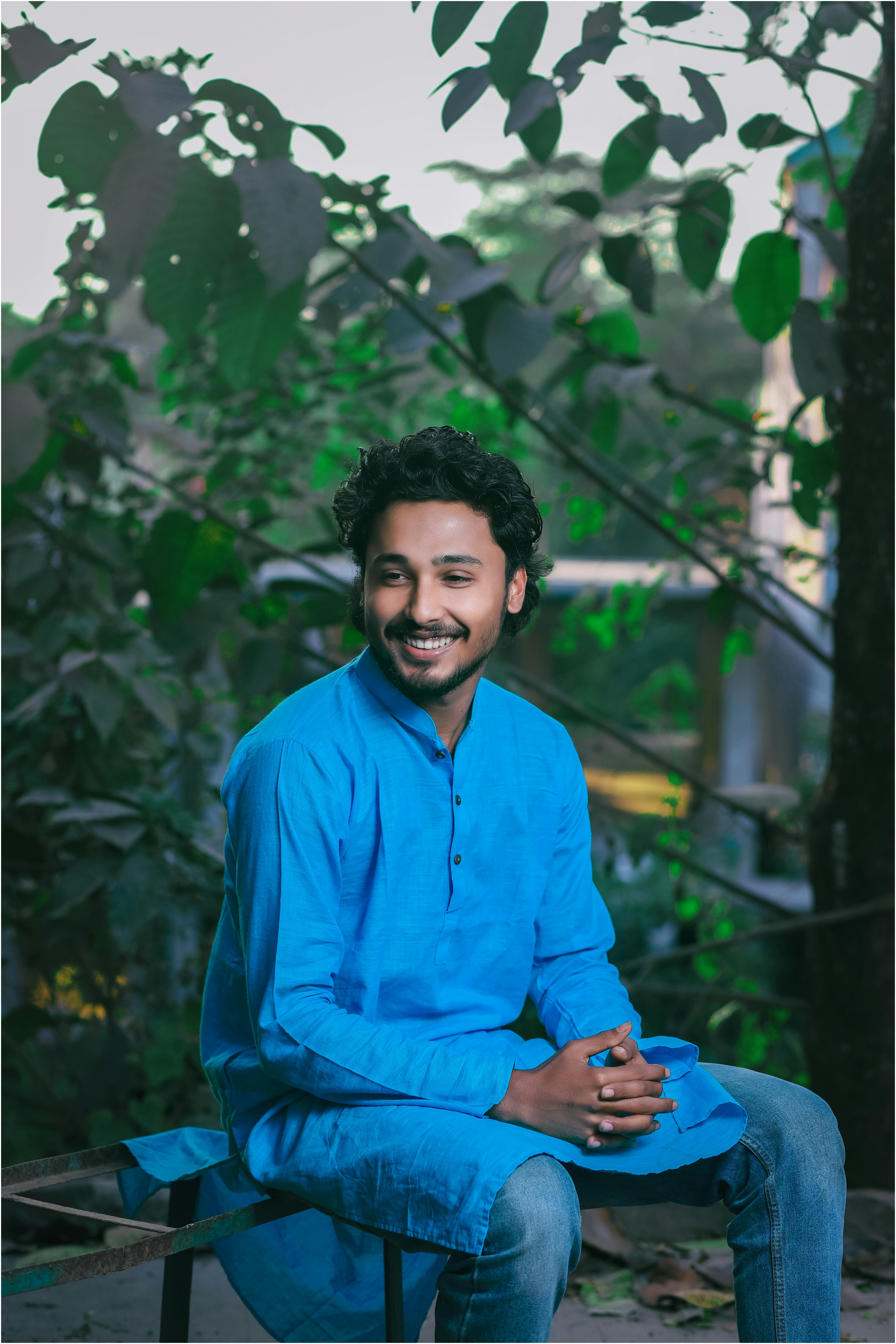 Portrait of a smiling young man in blue traditional attire, seated outdoors with greenery.