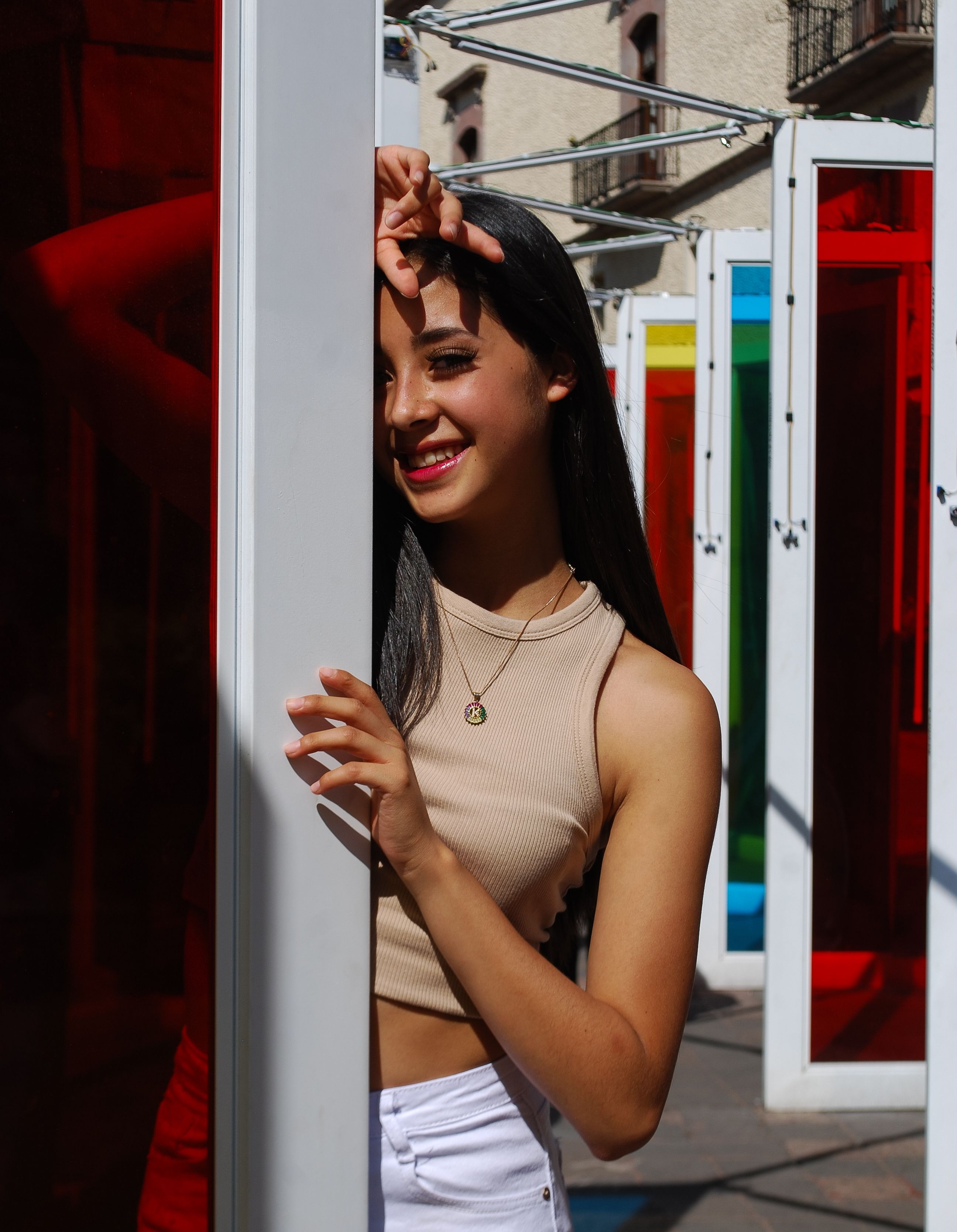 Fashionable woman posing with colorful architectural elements, smiling in sunlight.