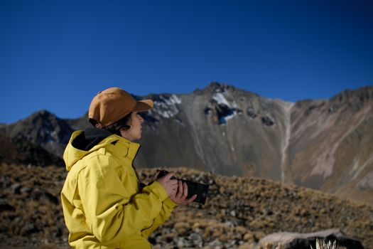 A woman in a yellow jacket and cap photographing stunning mountain landscapes on a clear day.