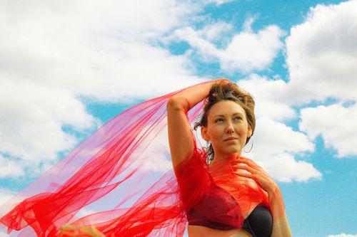 Free A woman poses gracefully with a red veil, set against a vibrant blue sky filled with clouds. Stock Photo