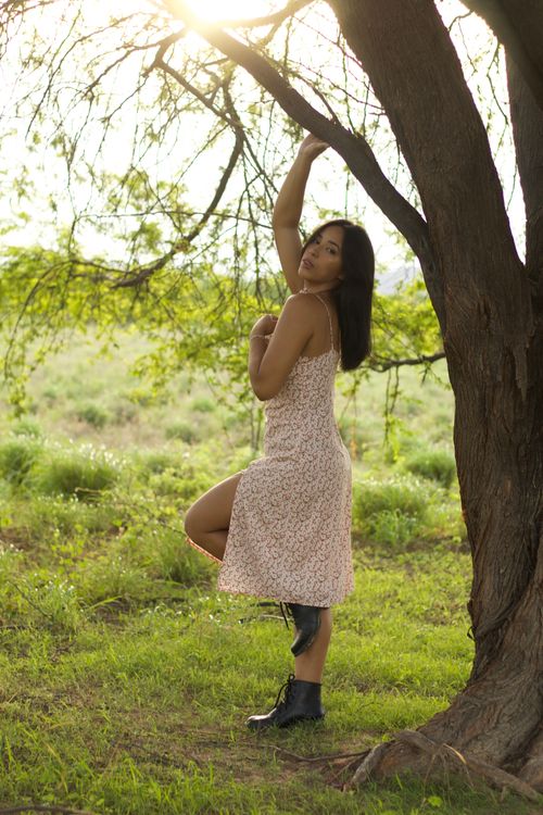 Free A woman in a floral dress poses elegantly under a tree in natural sunlight. Stock Photo