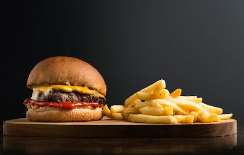 Appetizing burger with meat patty ketchup and cheese placed on wooden table with crispy french fries against black background