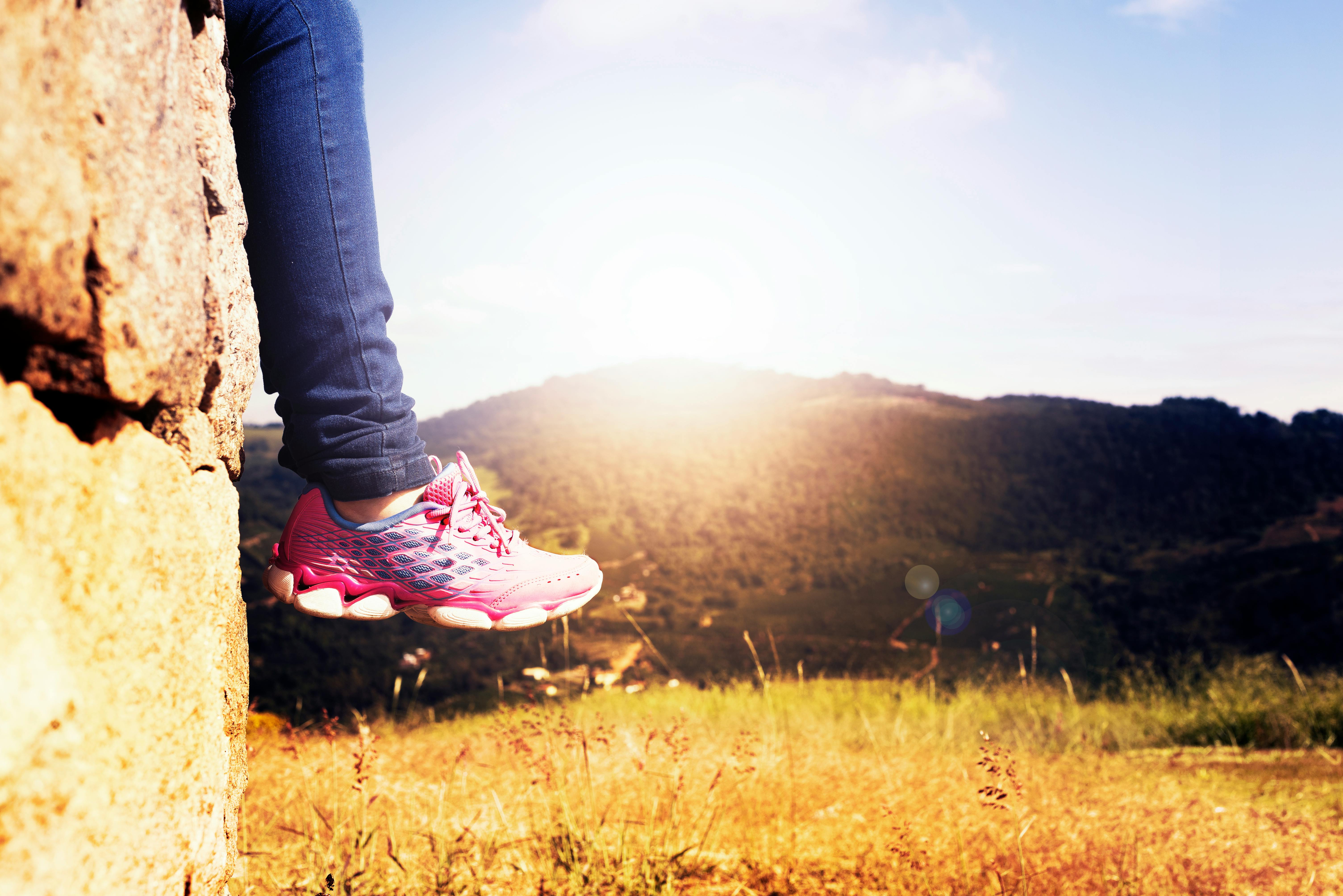 Person wearing pink sneakers sitting on a ledge overlooking a mountain landscape at sunset.