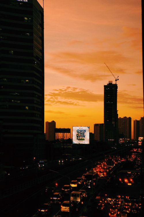 Free A bustling city street with skyscrapers and traffic at sunset, set against a vivid orange sky. Stock Photo