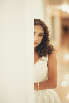 Bride in white lace dress subtly posing behind a curtain indoors.