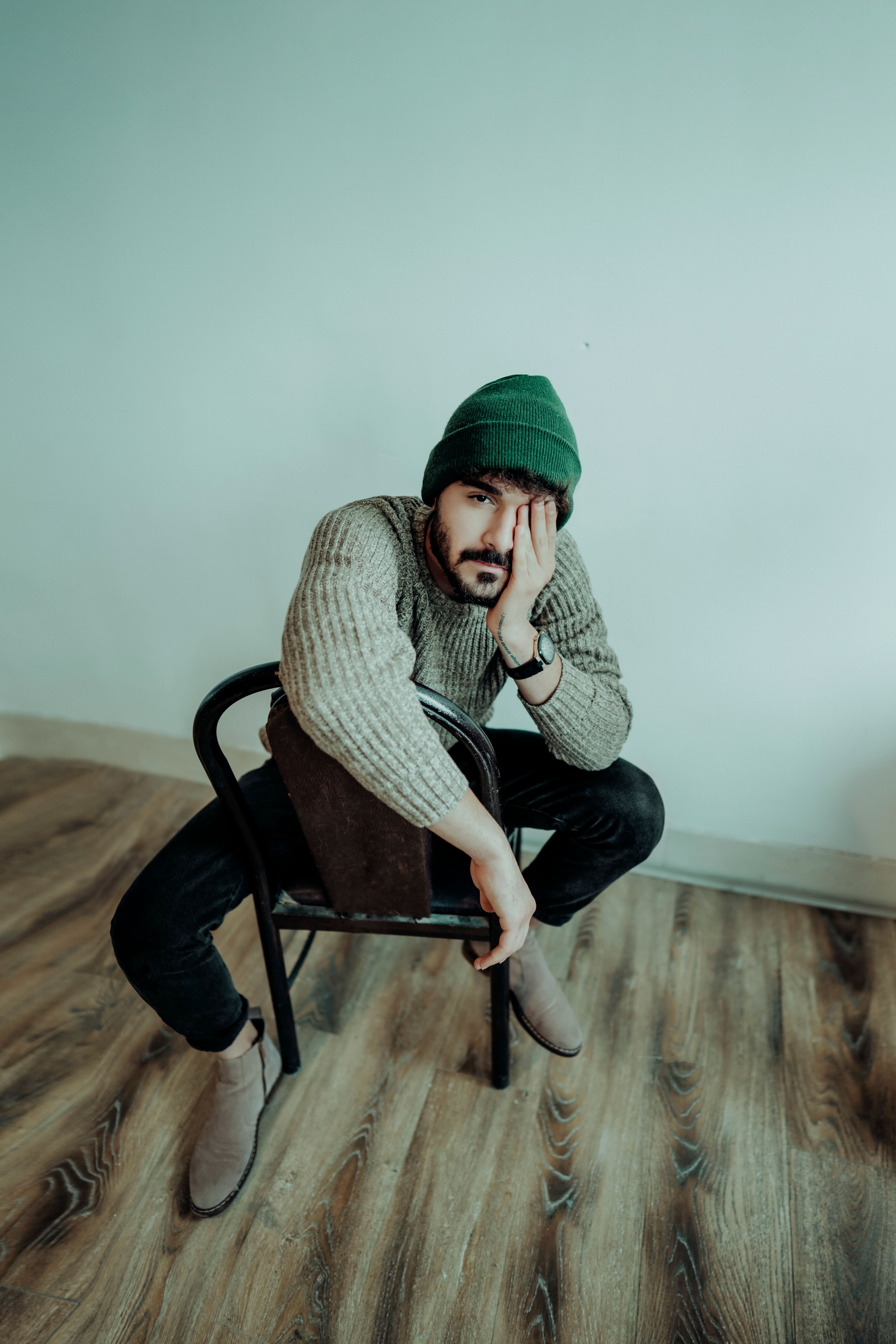 A man in casual attire poses thoughtfully on a wooden chair indoors, expressing style and introspection.