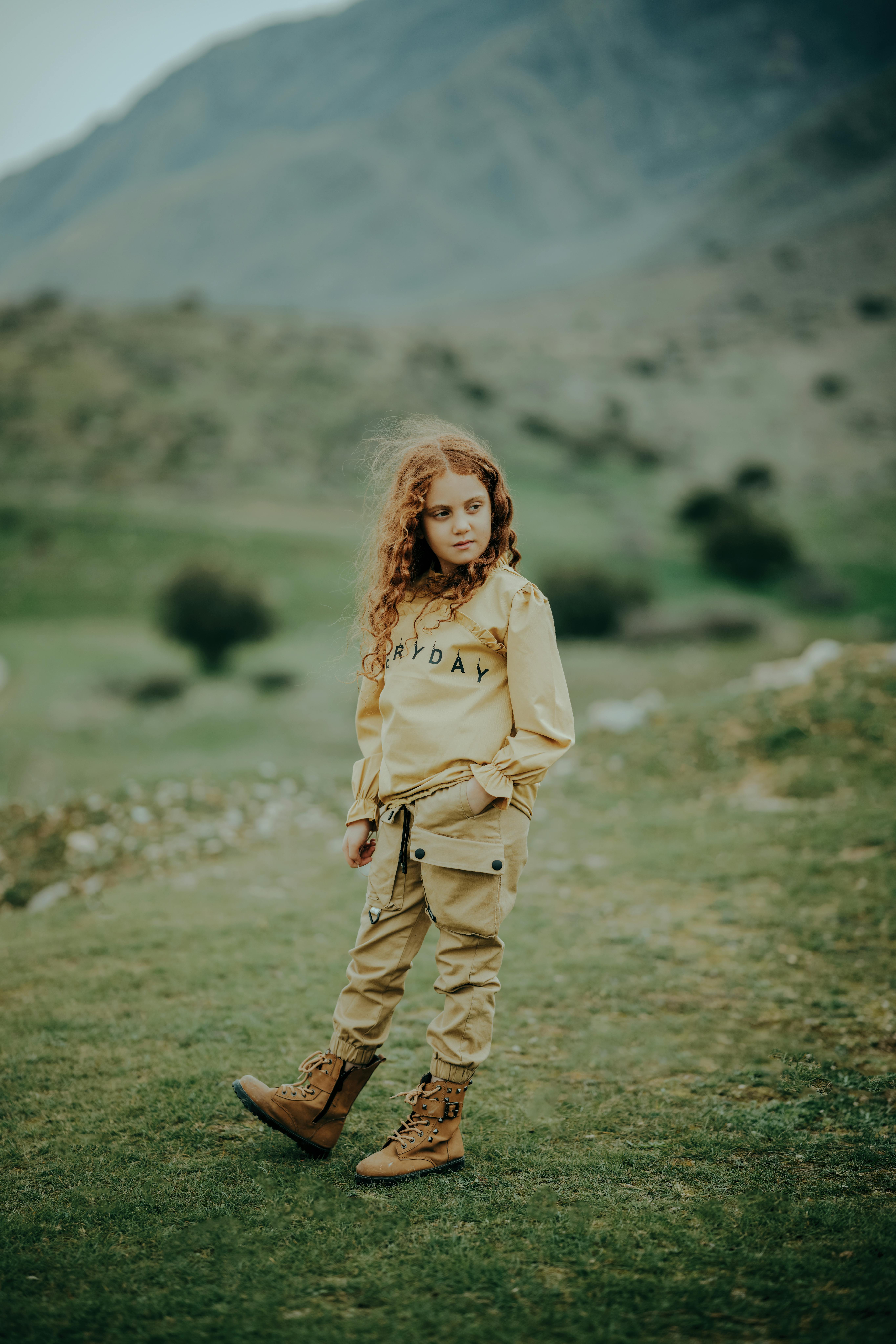 A young redhead girl poses fashionably in a rugged mountain field wearing a stylish outfit.