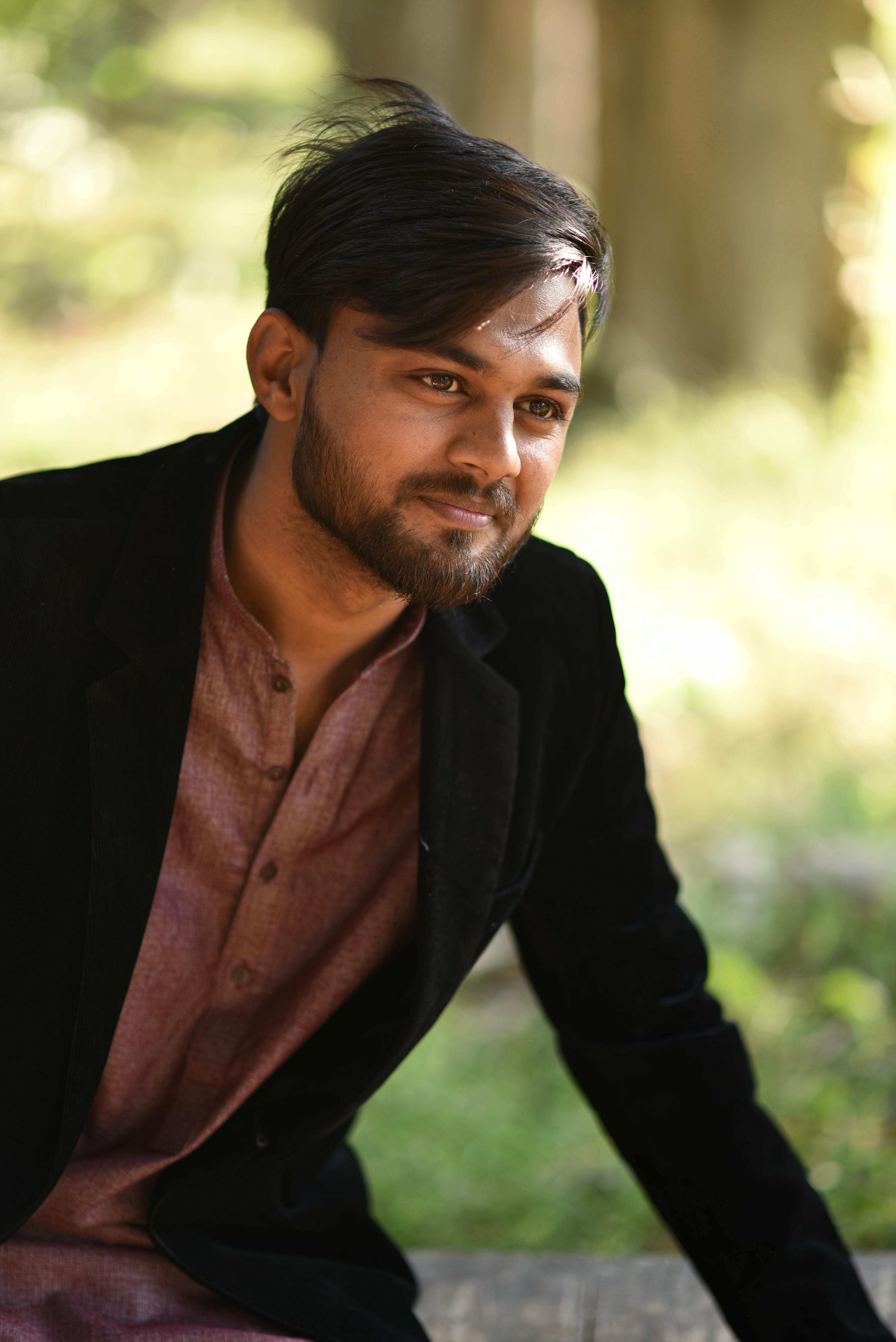 A stylish young man with black hair smiles outdoors, dressed in a velvet jacket.
