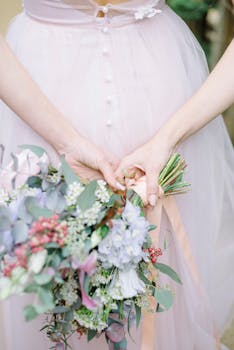 A close-up of hands holding a bouquet against a pink bridal gown, perfect for wedding themes.