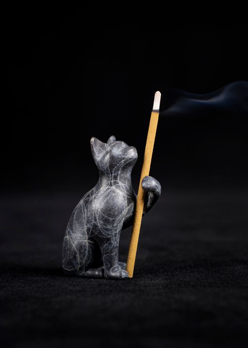 Free Marble cat figurine gracefully holding a burning incense stick, set against a dark background with visible smoke. Stock Photo