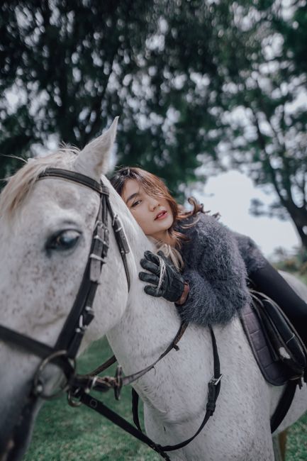 Stylish woman on horseback in natural setting, showcasing elegant fashion and thoughtful expression.