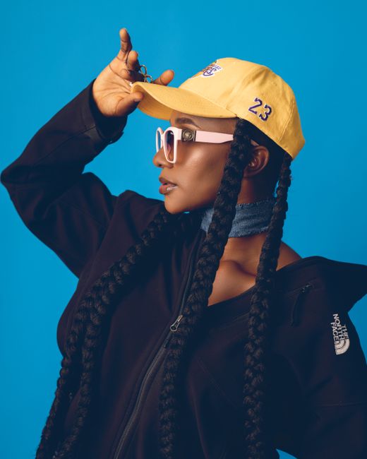 Fashionable woman wearing sunglasses and a jacket, striking a pose with a yellow cap in a studio setting.