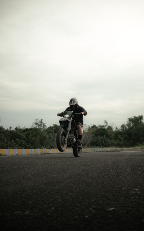 Free A motorcyclist performs an impressive wheelie on an open road under overcast skies, embodying adrenaline and skill. Stock Photo