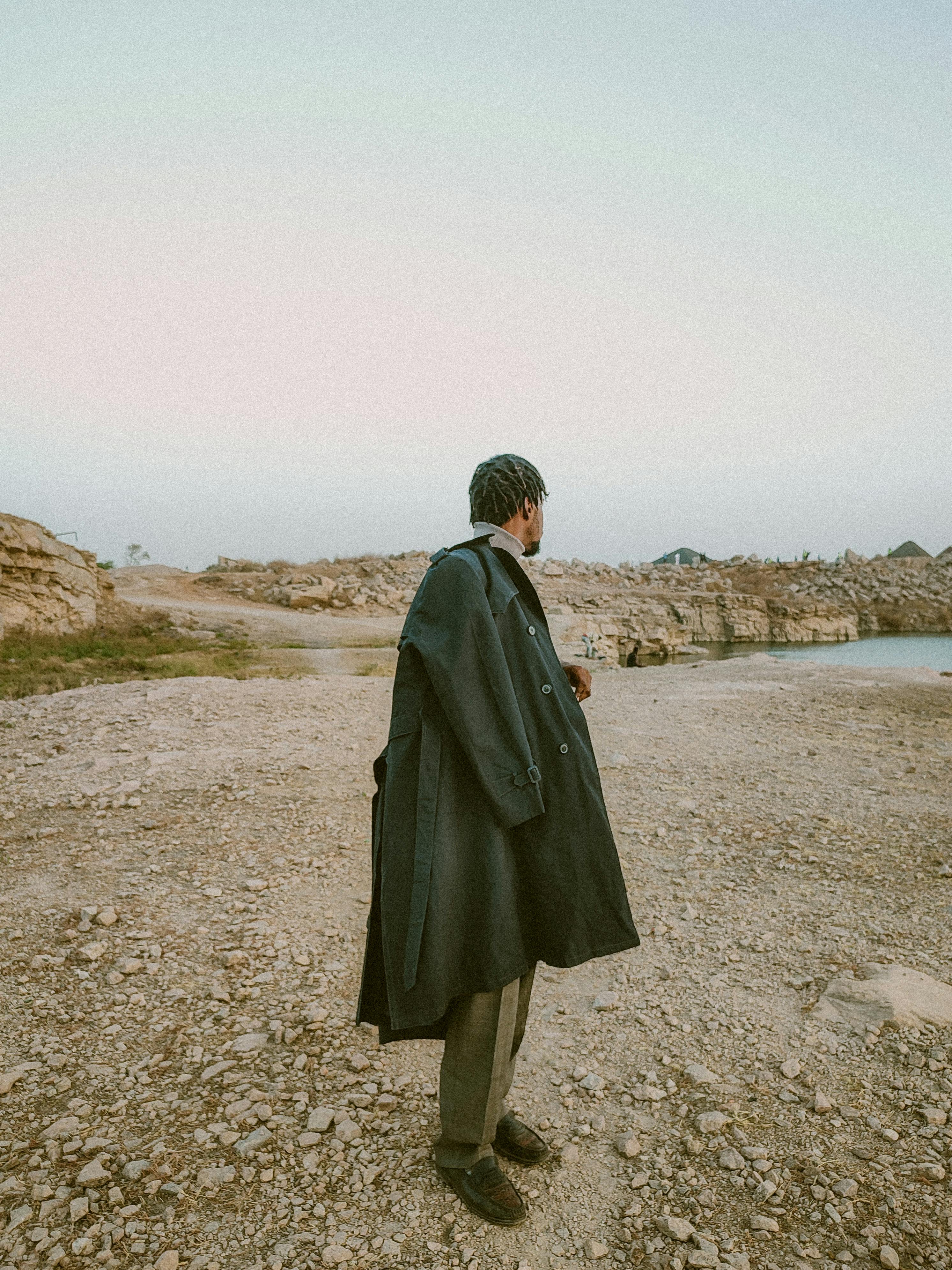 Man in a long coat standing on a rocky landscape under a clear sky.