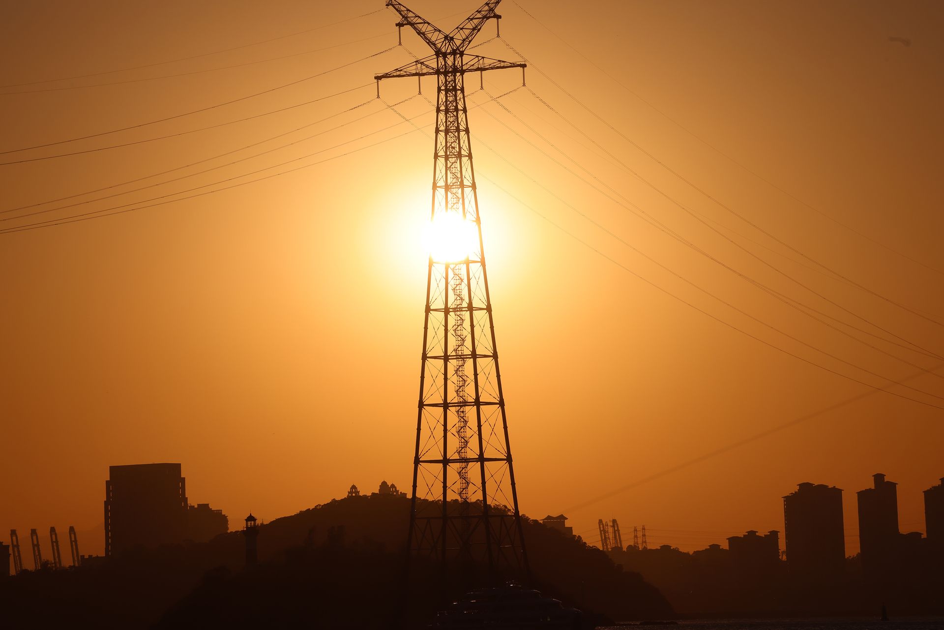 Silhouette of a transmission tower against an orange sunset sky in an urban setting.