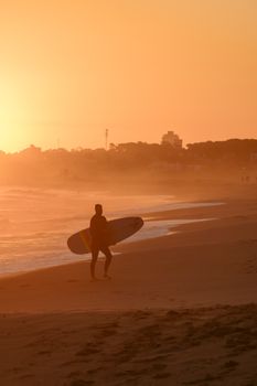 A surfer walks along Punta del Este beach at sunset, holding a surfboard against a warm, orange sky.