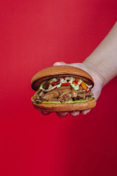 Close-up of a juicy beef burger held in hand with a vibrant red background. Perfect for fast food themes.