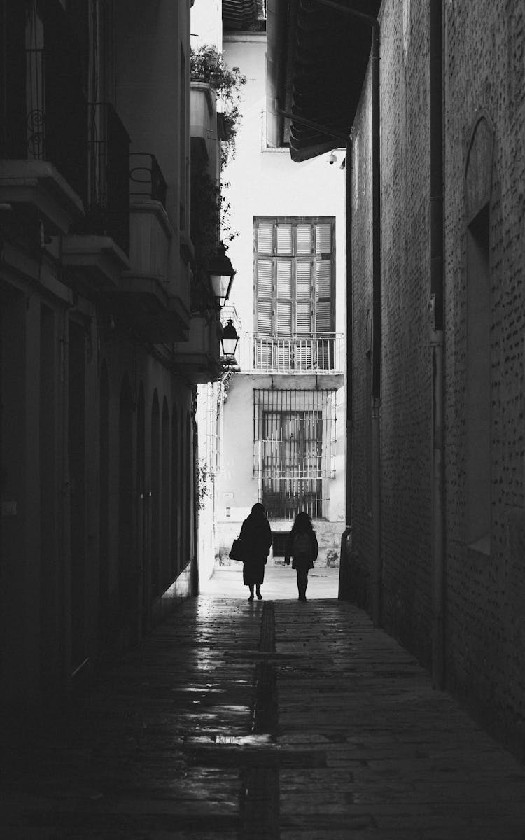 Silhouette Of Two People Walking In An Alley Between Buildings