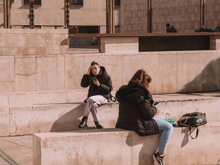 Dos mujeres sentadas en bancos de concreto en una plaza urbana soleada en Valencia, España, usando smartphones.