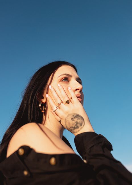 A young woman with tattoos poses against a clear blue sky, showcasing her unique style in Des Moines, IA.