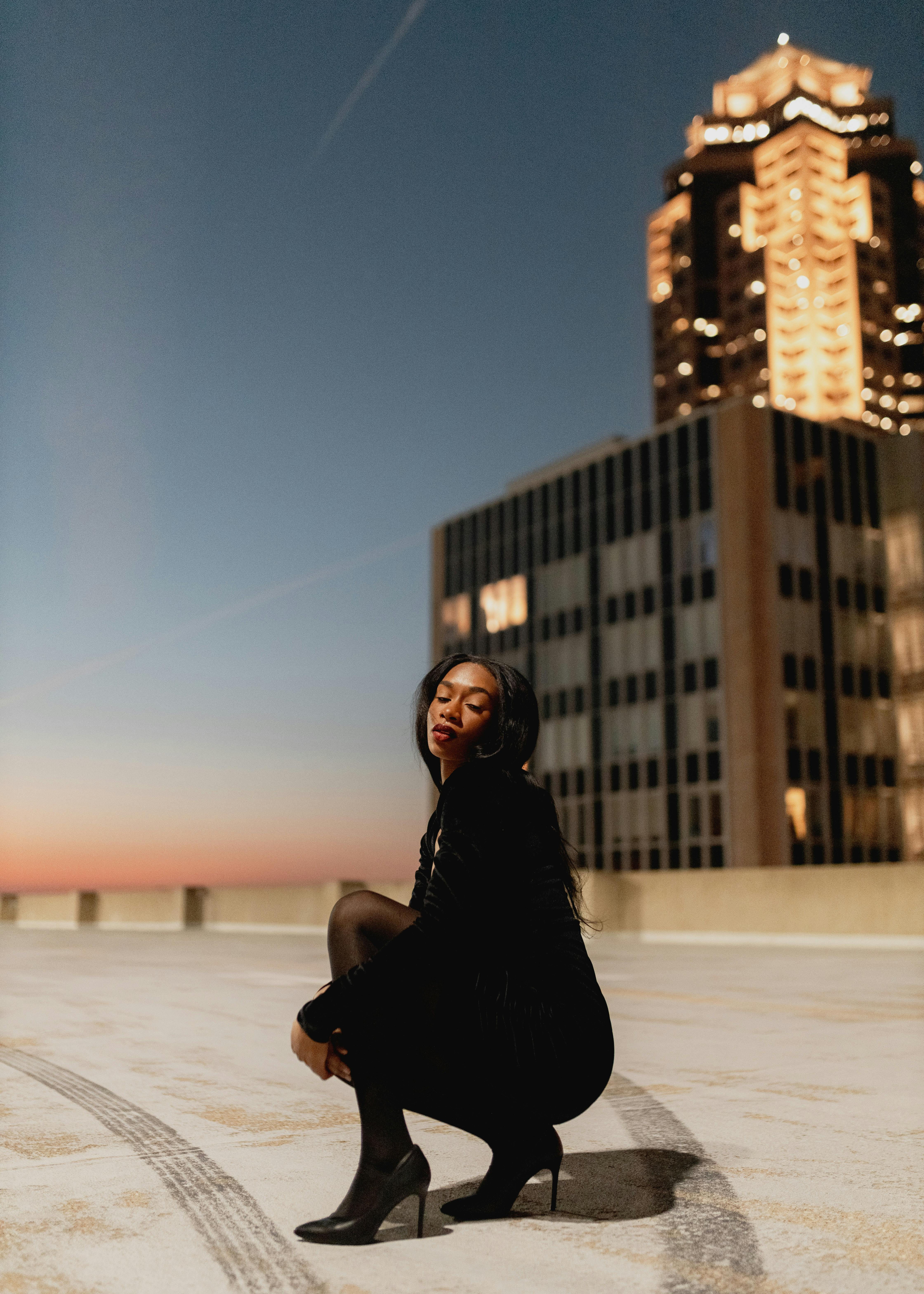 A woman in a black dress poses on a rooftop with city lights at twilight.