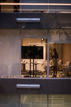 A woman stands by the window texting in a modern Moscow office at night.