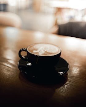 A warm cappuccino in a black cup with frothy latte art, set on a wooden table in a cozy cafe.