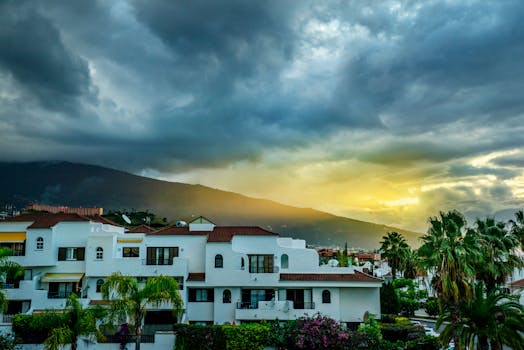 Vibrant sunset above tropical homes with lush palms and dramatic clouds.