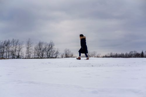 Free A woman in warm clothing walks across a snowy field under an overcast sky. Stock Photo