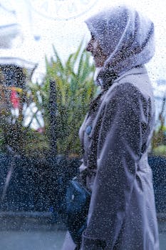 A woman in a coat stands behind a rain-soaked window, blending fashion with the beauty of weather.